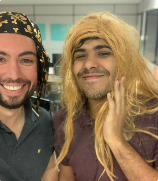 Two people smiling at the camera, one wearing a black bandana with a gold pattern and the other a blonde wig. Both are in an indoor setting, likely an office.