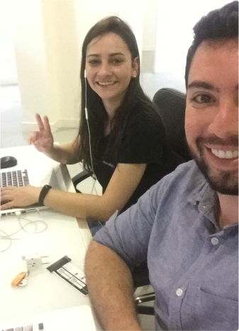 Two people sitting at a work desk, smiling at the camera. The person on the left is wearing earphones and making a peace sign with their fingers, while the person on the right is smiling broadly.