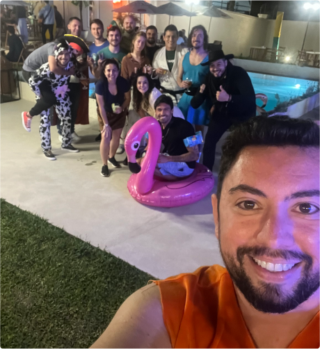 Um grupo de pessoas em um ambiente externo à noite, algumas vestindo fantasias. Eles estão posando para uma foto perto de uma piscina. Uma pessoa na frente está tirando a selfie, enquanto outros no fundo posam com uma boia rosa em formato de flamingo.