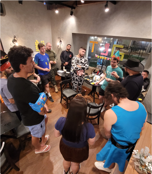 Um grupo de pessoas em uma sala, algumas usando fantasias. Elas estão reunidas em um círculo, conversando e interagindo. A sala é iluminada e tem uma janela grande ao fundo.