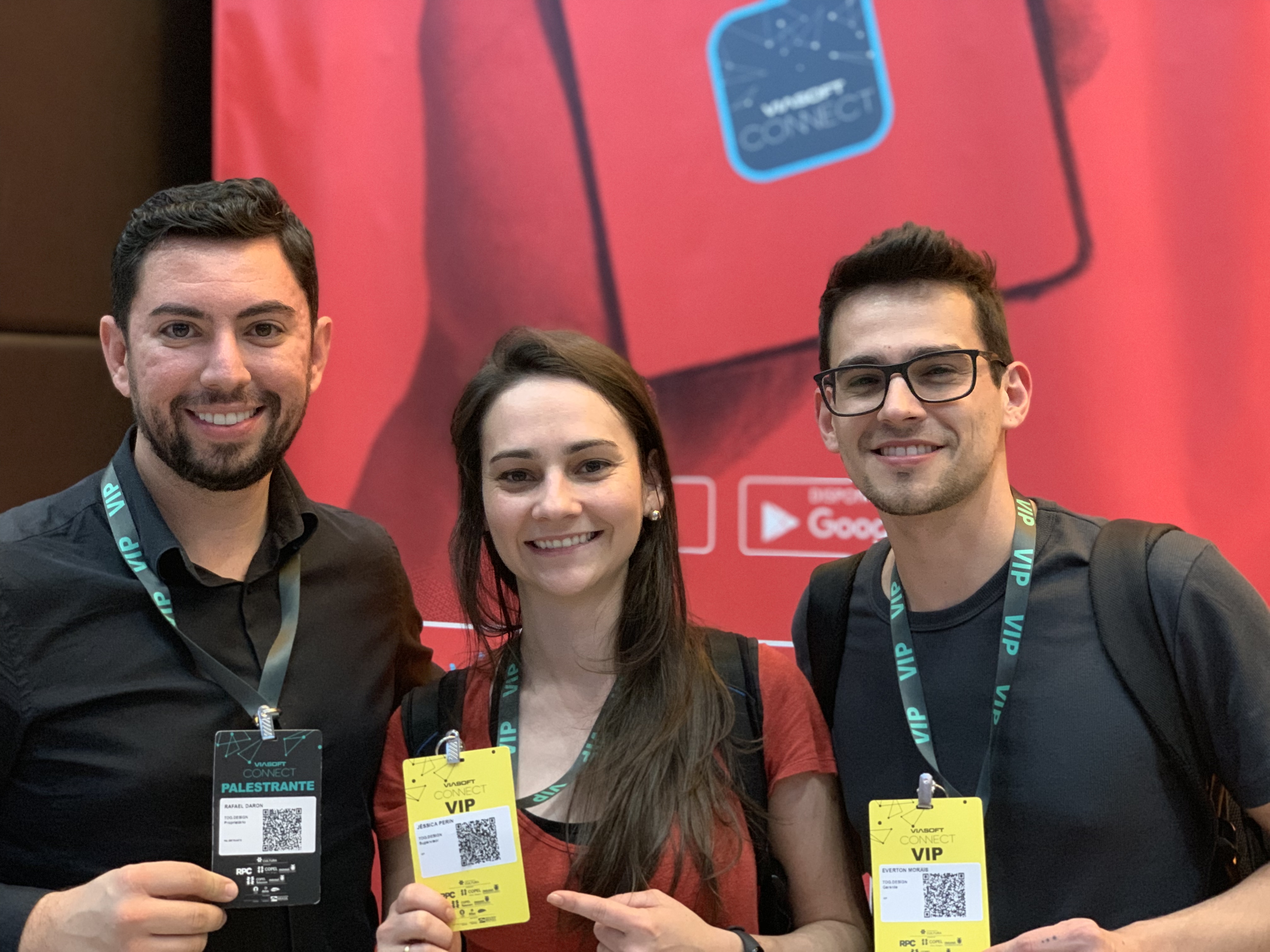 Três pessoas sorrindo, segurando crachás de identificação. A pessoa à esquerda é um palestrante, enquanto as outras duas têm crachás de VIP. O fundo é um banner vermelho de um evento.