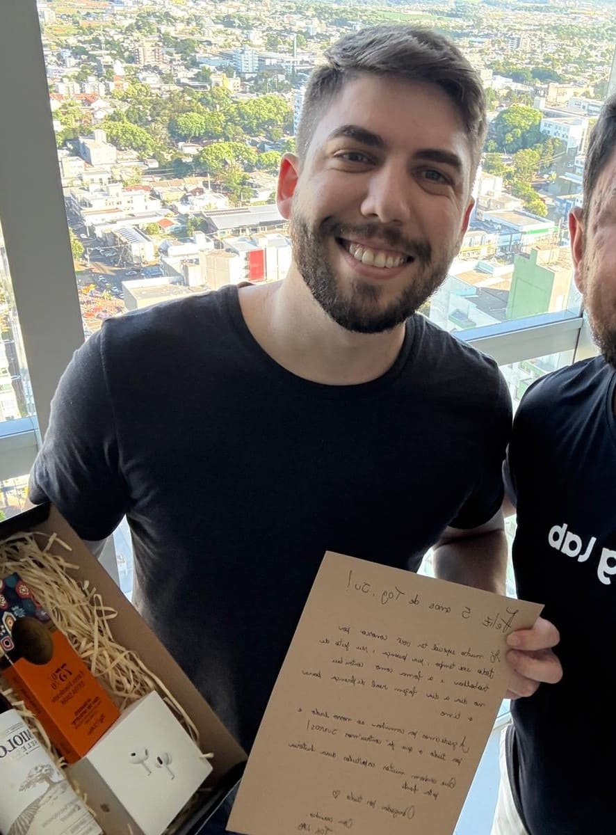 Toglab Recognition A smiling person holds a gift and a thank-you card, with a city view in the background from an indoor setting.