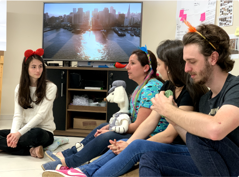 Grupo de quatro pessoas sentadas no chão de uma sala, algumas usando acessórios divertidos na cabeça. Ao fundo, há uma TV exibindo uma imagem de uma cidade ao pôr do sol.