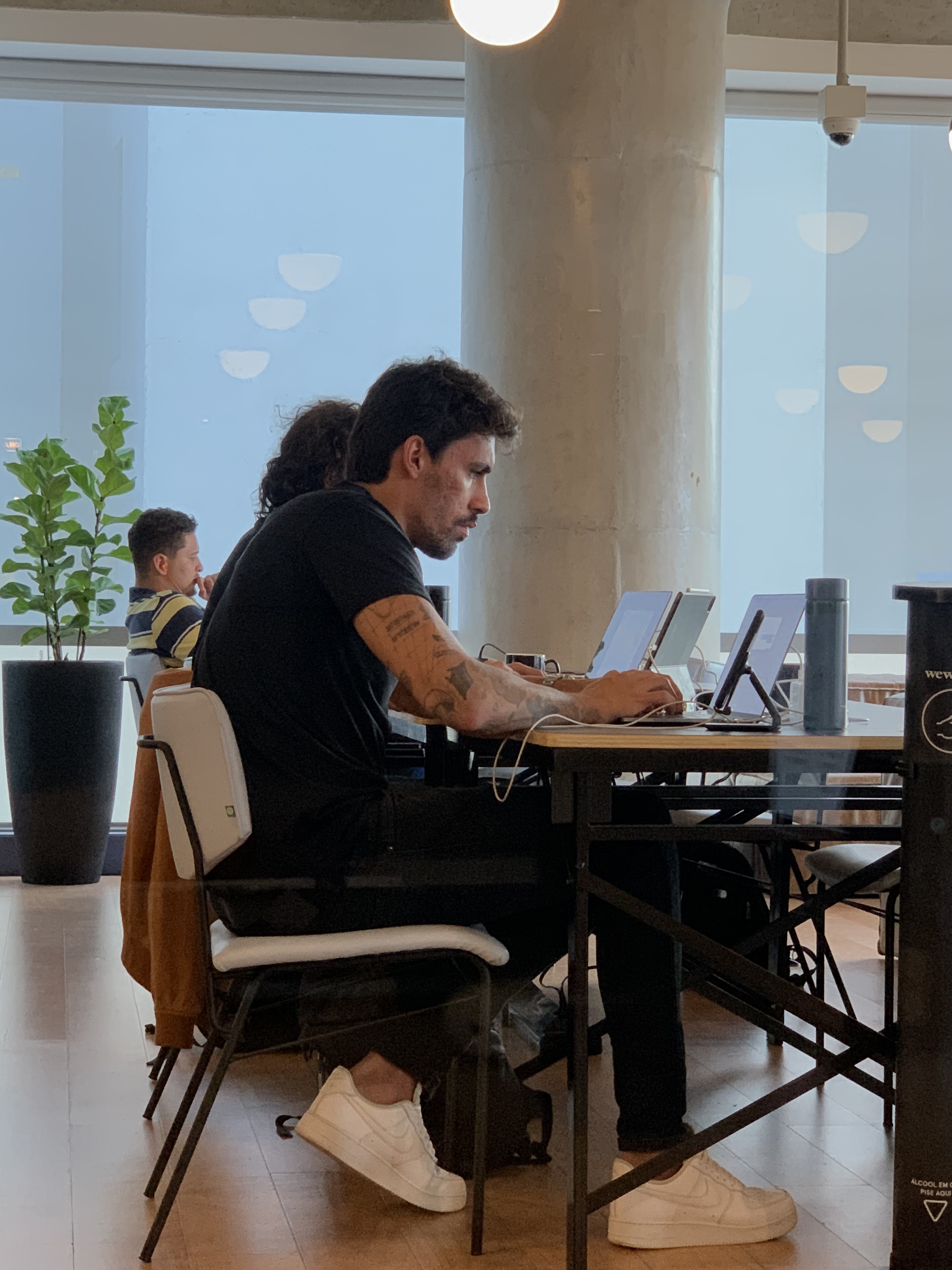 Homem trabalhando concentrado em um laptop em uma mesa de trabalho compartilhada. Há outras pessoas trabalhando ao fundo em um ambiente moderno com grandes janelas.