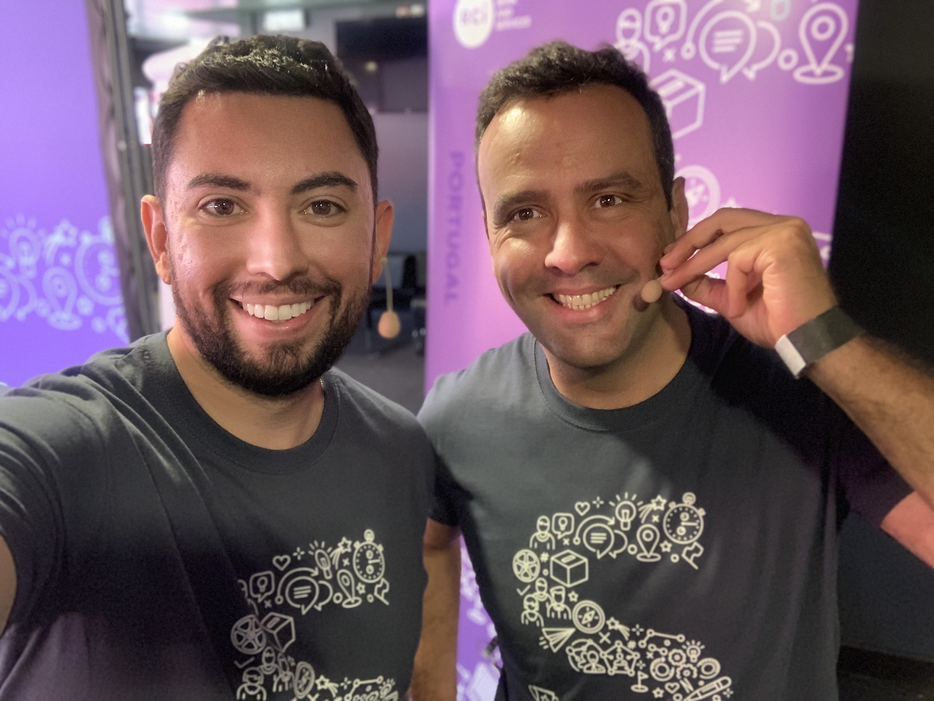 Dois homens sorrindo e tirando uma selfie. Ambos estão usando camisetas pretas com um design gráfico e um deles está usando um microfone de headset. O fundo é decorado com gráficos em roxo.