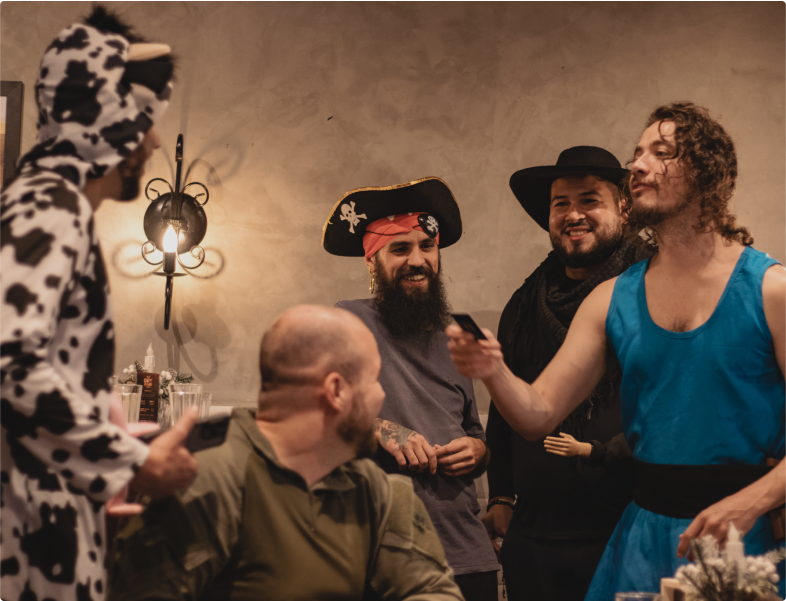 Five people in an indoor setting, all dressed in costumes. One person is in a cow costume, another as a pirate, another with a cowboy hat, and another in a blue costume. They are smiling and talking, creating a cheerful and relaxed atmosphere.