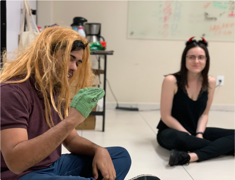 Duas pessoas sentadas no chão em um ambiente interno. A pessoa à esquerda está usando uma peruca loira e uma luva verde, enquanto a pessoa à direita está vestindo uma tiara com chifres vermelhos. Ambas estão sorrindo e parecem estar se divertindo.