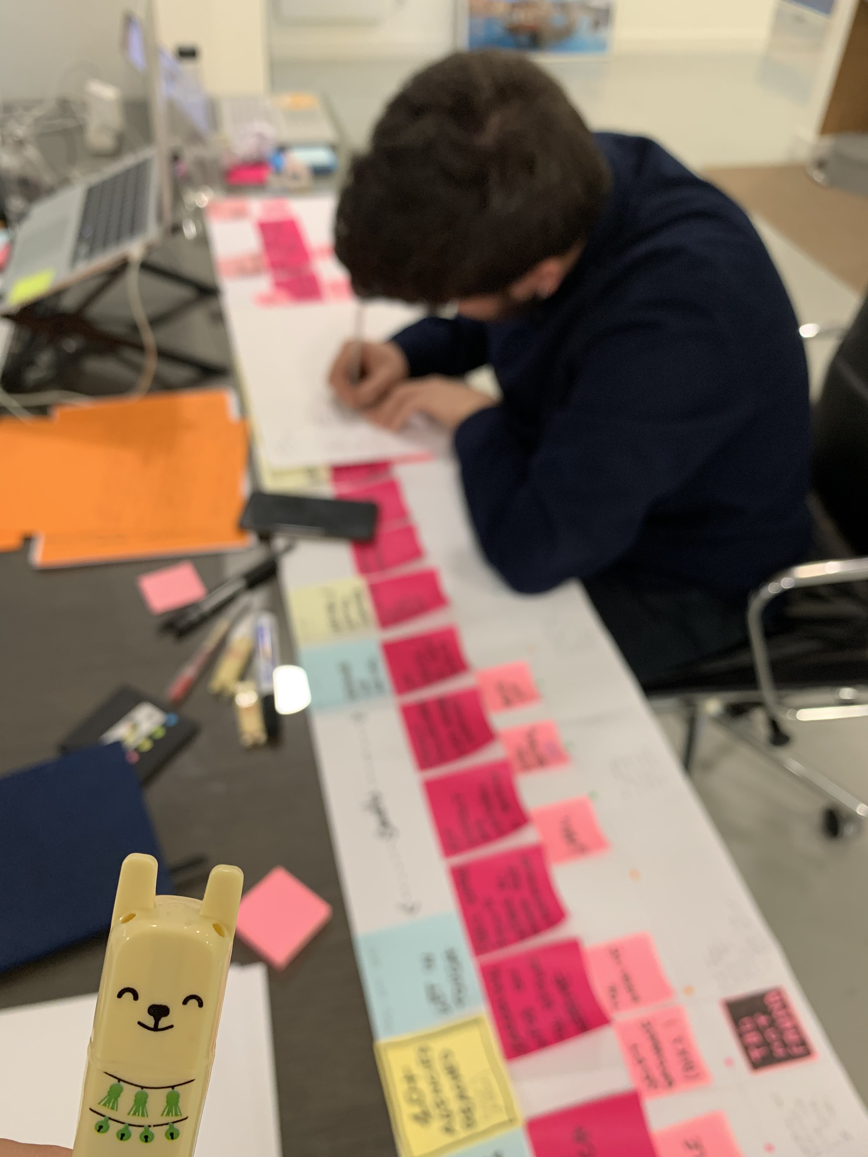 Person writing at a table filled with colorful post-it notes organized in a line. In the foreground, there is an animal-shaped paper clip.