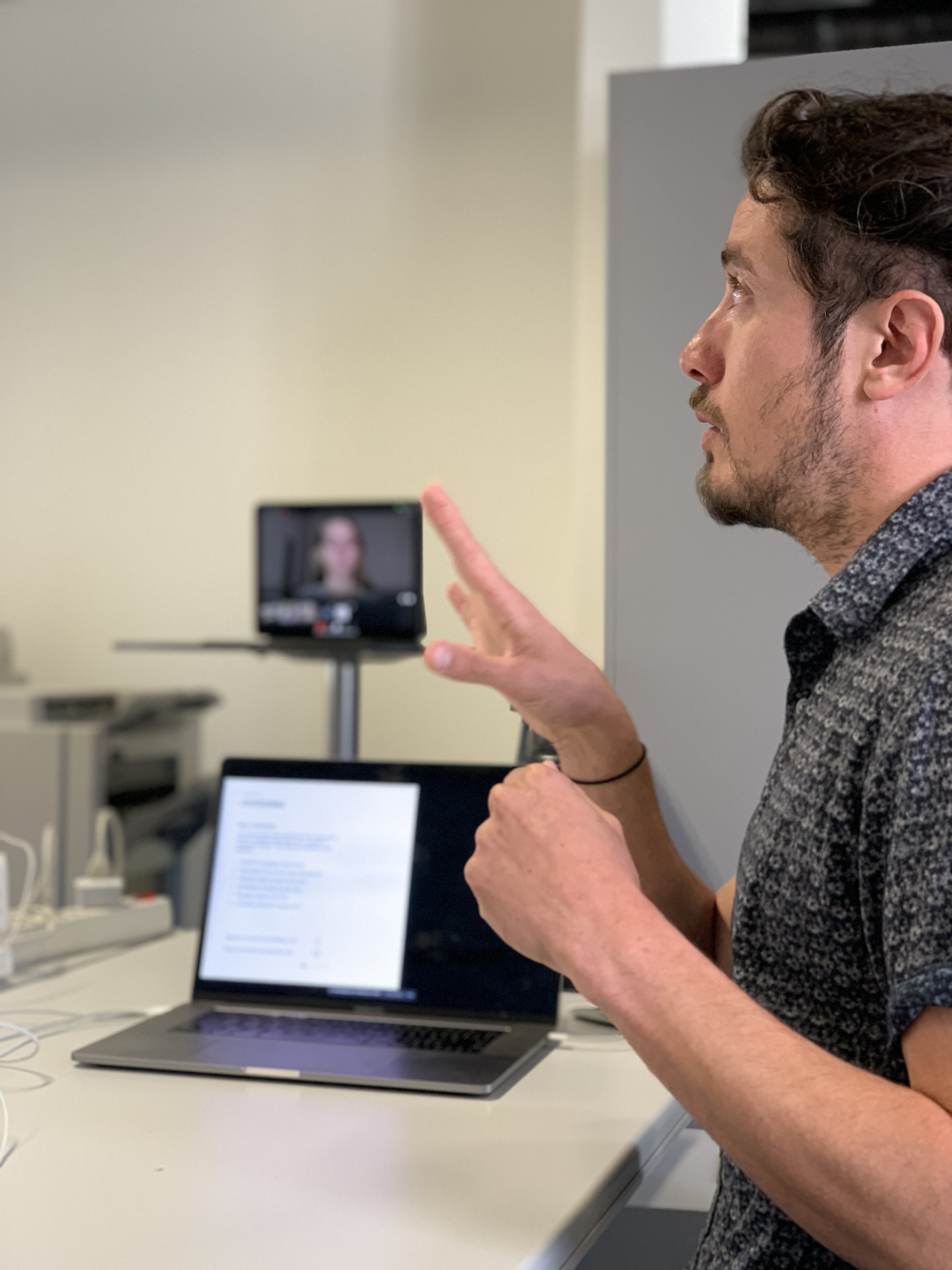 Homem gesticulando enquanto faz uma apresentação, com um laptop aberto à sua frente e uma pessoa em uma videochamada visível ao fundo na tela.