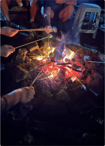 Várias pessoas assando marshmallows em um fogo de acampamento à noite. As mãos seguram espetos com marshmallows sobre as chamas, criando um clima acolhedor e amigável.