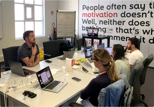 Quatro pessoas sentadas ao redor de uma mesa de reunião, cada uma com um laptop, participando de uma reunião sobre design. Uma pessoa está apresentando algo, e há uma lousa e um monitor com uma videochamada ao fundo. A parede possui uma citação motivacional.