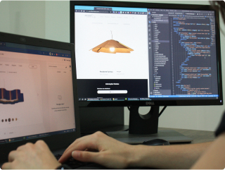 Person working on a laptop and a monitor. The monitor displays a product design website and programming code.