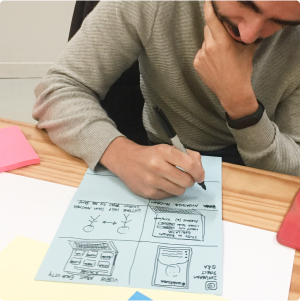 Person drawing a wireframe on a blue sheet of paper at a table. Around the table, there are colorful post-it notes and other stationery materials.