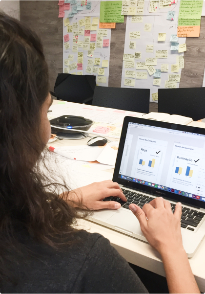 Person working on a laptop at an office table. The laptop displays a prototype in Figma. The background wall is covered with colorful post-it notes with annotations.