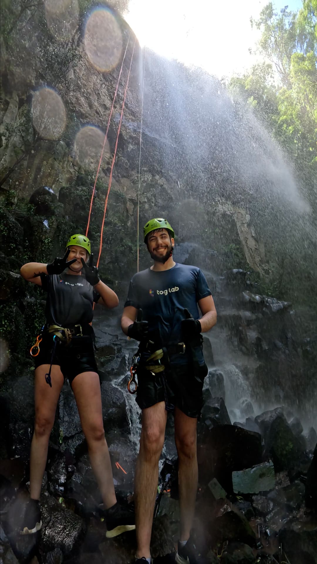 Two colleagues smiling at the camera. wo people wearing helmets and rappelling gear smile while posing under a waterfall in a natural setting.