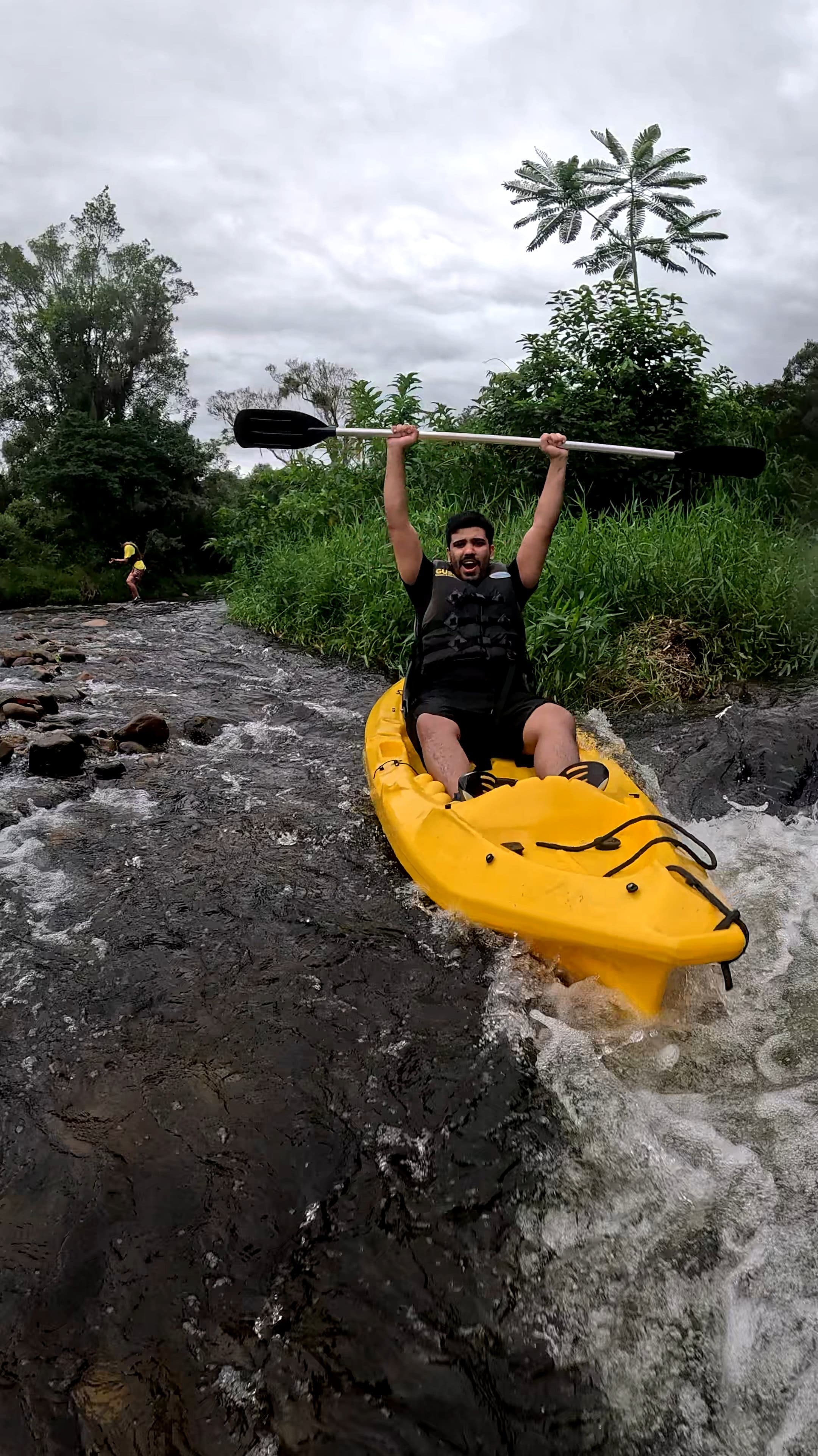Team member kayaking through a river rapid. Team member wearing a life jacket kayaking down a shallow rocky river with paddle raised, surrounded by green vegetation.