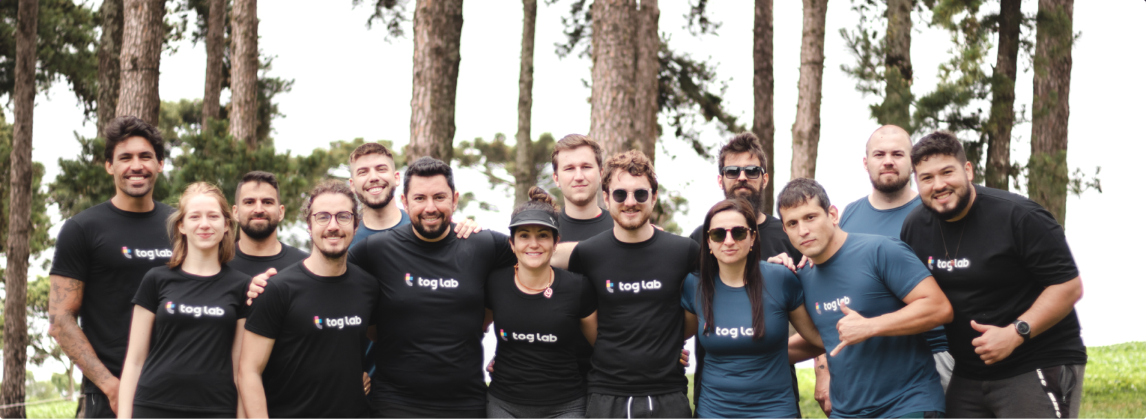 Um grupo de quinze pessoas, todas usando camisetas pretas com o logotipo tog lab, posando juntas ao ar livre, em um ambiente arborizado.