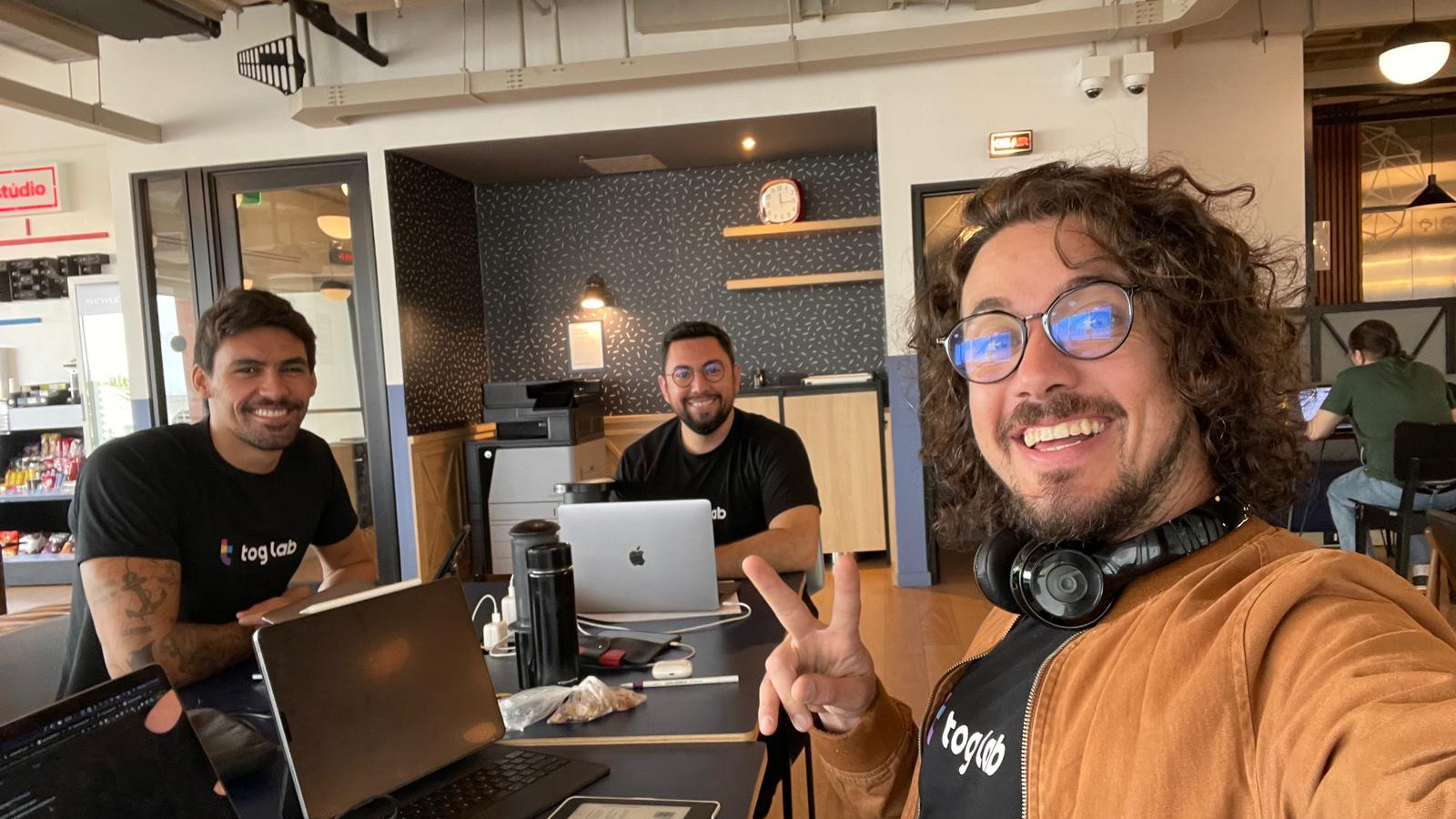 Três homens sorrindo e trabalhando em laptops em um ambiente de escritório. Dois estão sentados à mesa e um está tirando a selfie. Todos estão vestindo camisetas com o logo tog lab.