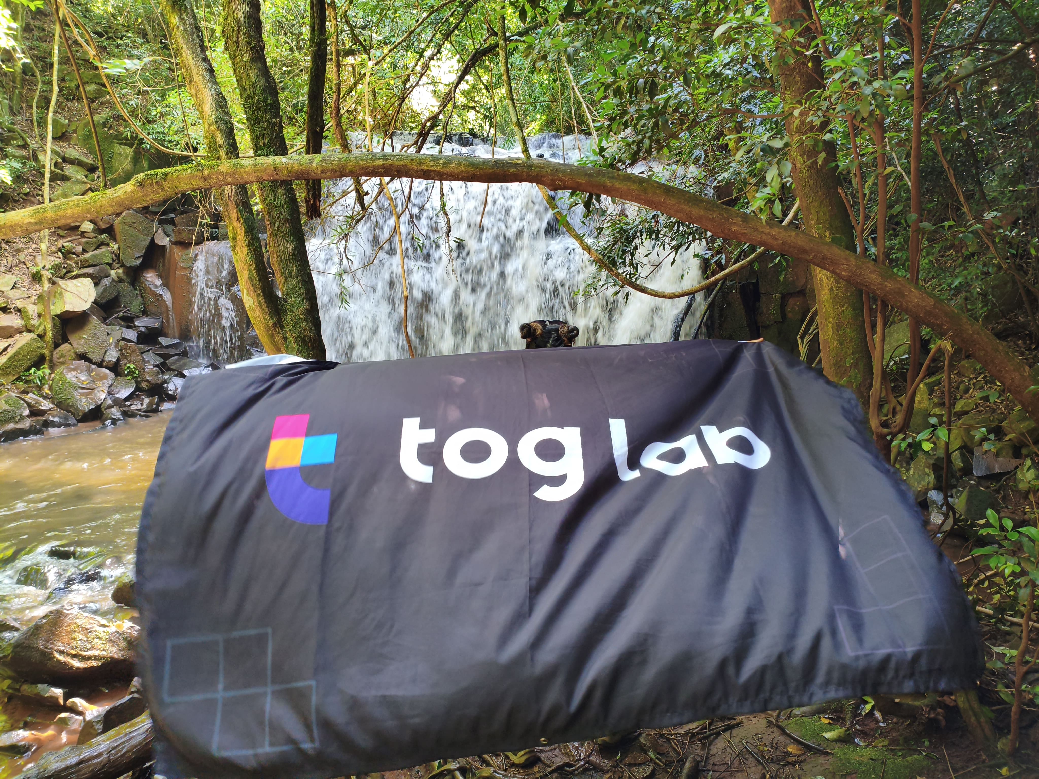 Toglab in nature. A Toglab banner is displayed in front of a waterfall, surrounded by lush vegetation in a natural setting.