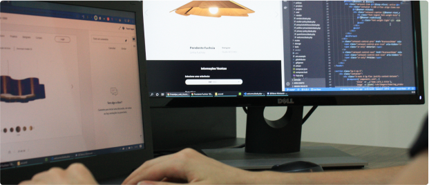 Pessoa trabalhando em um laptop e monitor. O monitor à direita exibe código de programação, enquanto o laptop à esquerda mostra um site de design de produto com uma luminária.